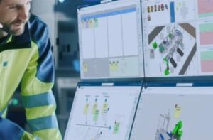 A factory worker in a high-visibility jacket is looking at multiple computer monitors displaying 3DEXPERIENCE software.