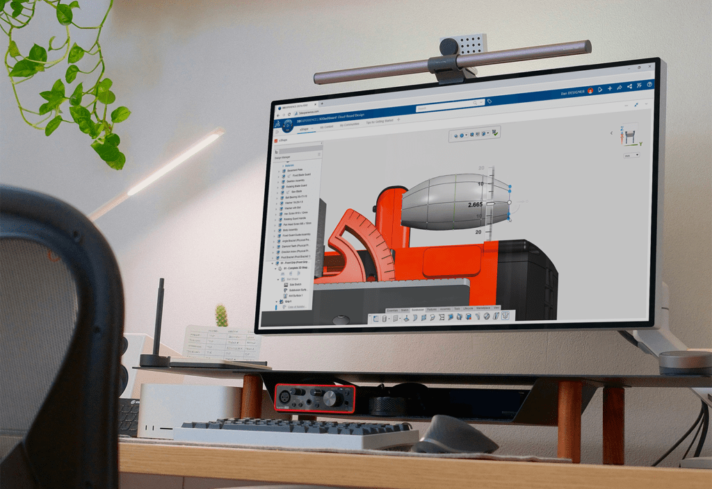 A modern workspace with a large monitor displaying a 3DEXPERIENCE platform interface, showing a 3D CAD model in design mode. The desk setup includes a keyboard, mouse, and a clean, minimalistic environment.